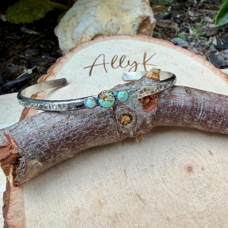 May include: A silver cuff bracelet with a hammered texture and four turquoise stones. The bracelet is displayed on a wooden surface with the handwritten text "Ally K". The turquoise stones are a mix of blue and brown.