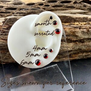 May include: A white ear-shaped display stand featuring six round red gemstone stud earrings. The text labels indicate smooth and serrated edge styles, with sizes 4mm, 5mm, and 6mm shown for reference on the display.