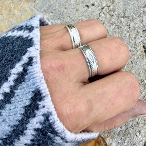 May include: Two silver-toned metal rings worn on adjacent fingers. Each ring features a wide band with a textured, grooved edge and a central panel displaying a repeating geometric diamond-shaped pattern engraved into the metallic surface.