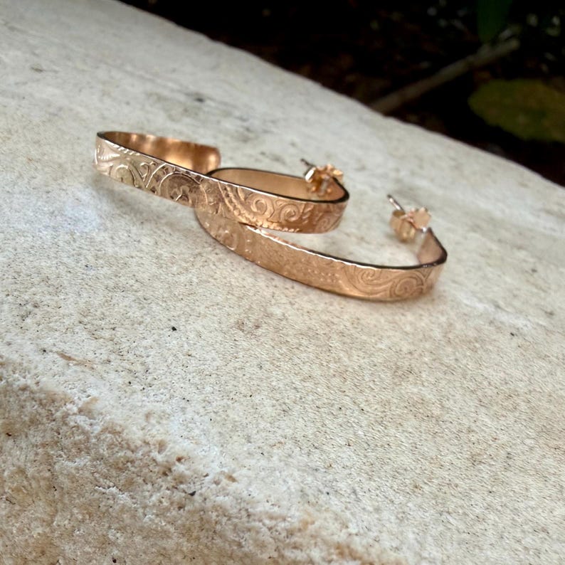 May include: A pair of gold-colored hoop earrings with an engraved floral pattern. The earrings are open at the bottom and have a post closure. The earrings are displayed on a light-colored, textured surface.