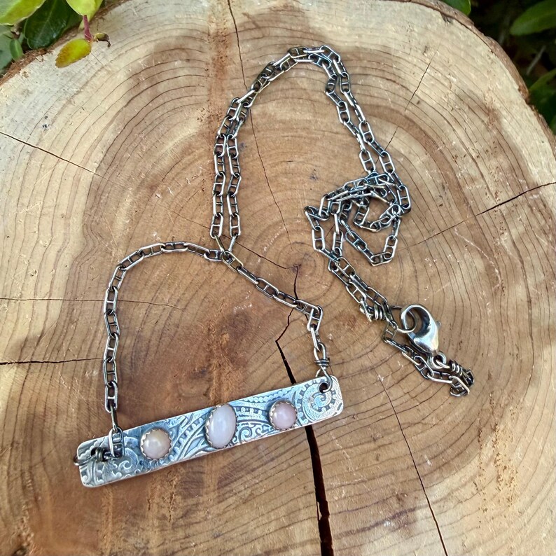 May include: A silver-toned metal necklace featuring a rectangular bar pendant with an intricate etched scrollwork pattern. The bar is set with three smooth, oval-shaped, pale pink cabochon stones. The pendant hangs from a sturdy link chain.