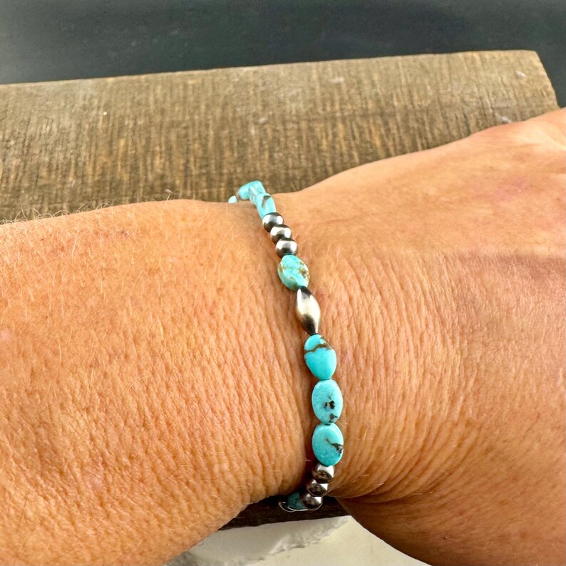 May include: A turquoise and silver beaded bracelet. The bracelet features oval turquoise stones interspersed with small silver beads and elongated silver beads. The bracelet is worn on a wrist.