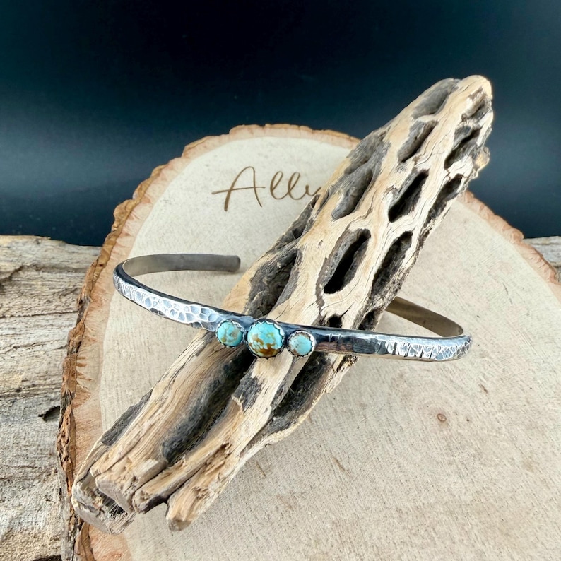 May include: A silver cuff bracelet with a hammered texture and three turquoise stones. The bracelet is displayed on a piece of weathered wood. The word "Ally" is written on the wood.