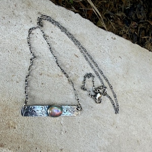 May include: A silver-toned necklace featuring a rectangular metal bar pendant with an intricate embossed scroll pattern. An oval-shaped cabochon stone with iridescent, multi-colored reflections is set in the center of the bar.