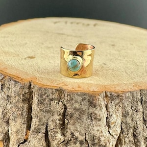 May include: A gold-colored ring with a turquoise stone set in the center. The ring has a hammered texture and a wide band. The ring is displayed on a wooden surface.