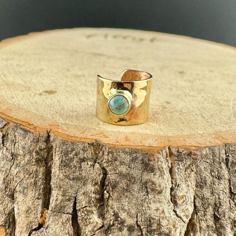May include: A gold-colored ring with a turquoise stone set in the center. The ring has a hammered texture and a wide band. The ring is displayed on a wooden surface.