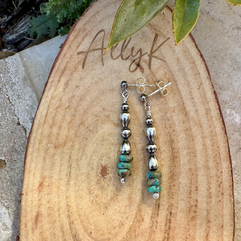 May include: A pair of silver earrings with a dangle design. Each earring features a series of silver beads and turquoise-colored stones. The earrings are displayed on a wooden surface with the handwritten text "Adly K".