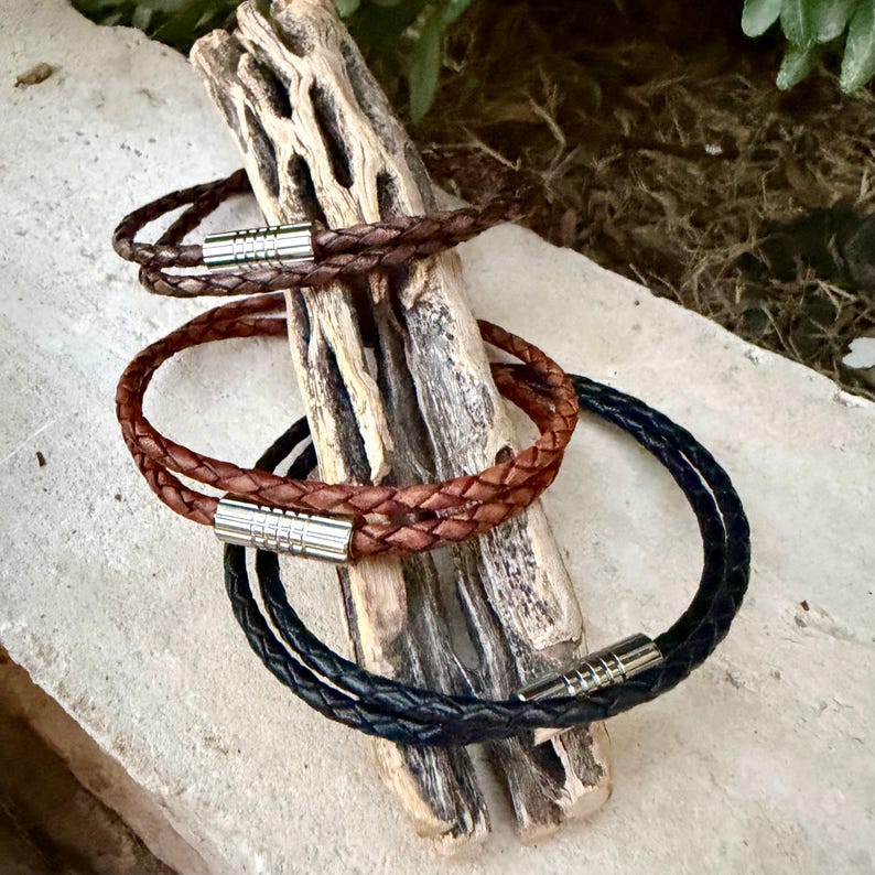 May include: Three braided leather bracelets in brown, tan, and black, each featuring a polished silver-tone cylindrical magnetic clasp. The bracelets are displayed stacked on a piece of textured, light-colored weathered wood.