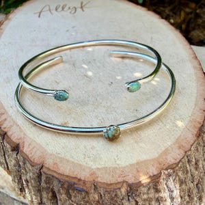 May include: Two silver-toned cuff bracelets with turquoise-colored stones. One bracelet is a double band design, and the other is a single band. The bracelets are displayed on a wooden surface with the handwritten name "Allyk".