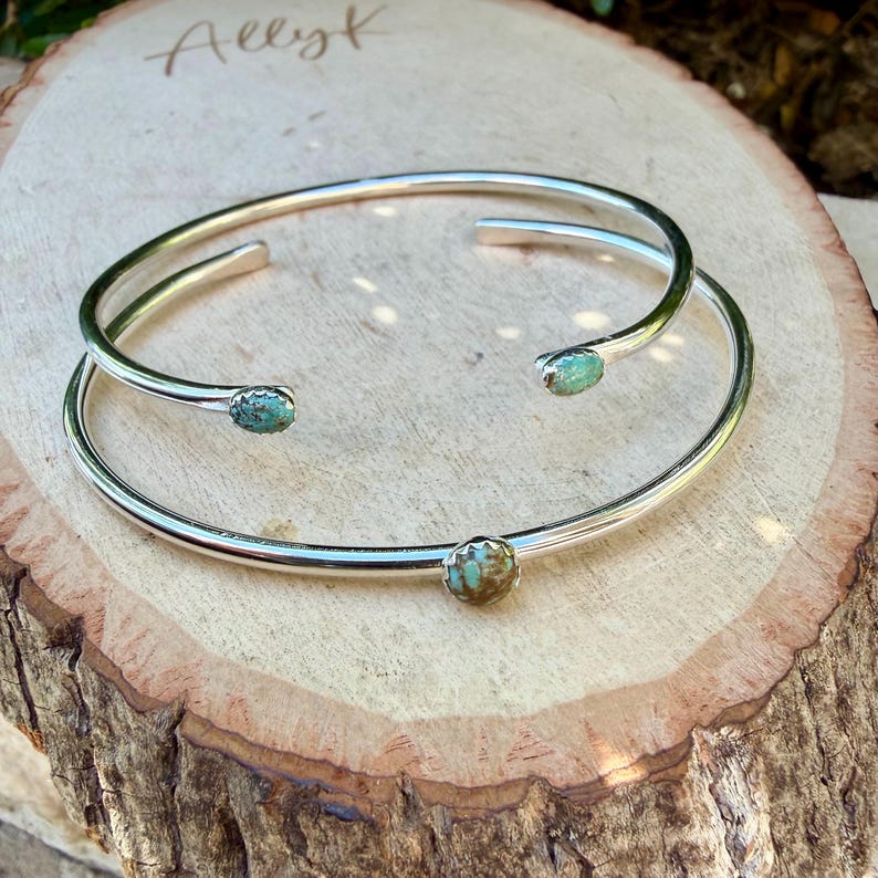 May include: Two silver-toned cuff bracelets with turquoise-colored stones. One bracelet is a double band design, and the other is a single band. The bracelets are displayed on a wooden surface with the handwritten name "Allyk".