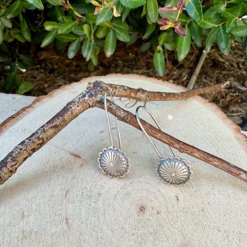 May include: A pair of silver-toned drop earrings featuring circular, sunburst-patterned medallions with a central dot. The earrings have long, curved wire hooks and are suspended from a thin, textured brown branch.