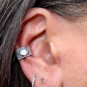 May include: A silver ear cuff with a textured, bark-like design. The cuff features a round, iridescent stone. A small, clear stud earring and a small hoop earring are also visible.