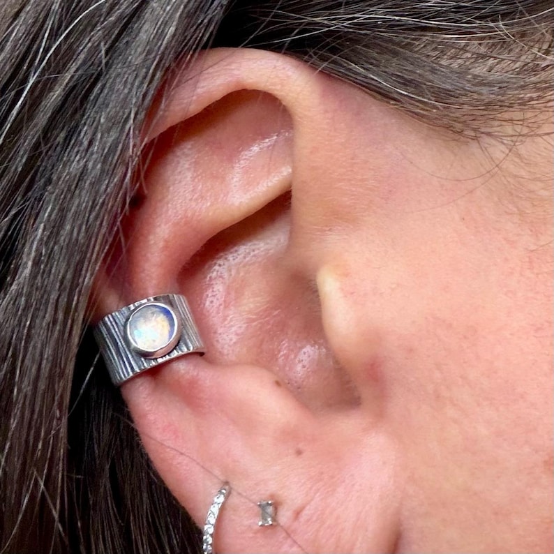 K&ouml;nnte beinhalten: Ein silberner Ohrklemme mit einem strukturierten, rindenartigen Design. Die Manschette hat einen runden, irisierenden Stein. Ein kleiner Ohrstecker und ein kleiner Creolen-Ohrring sind ebenfalls sichtbar.