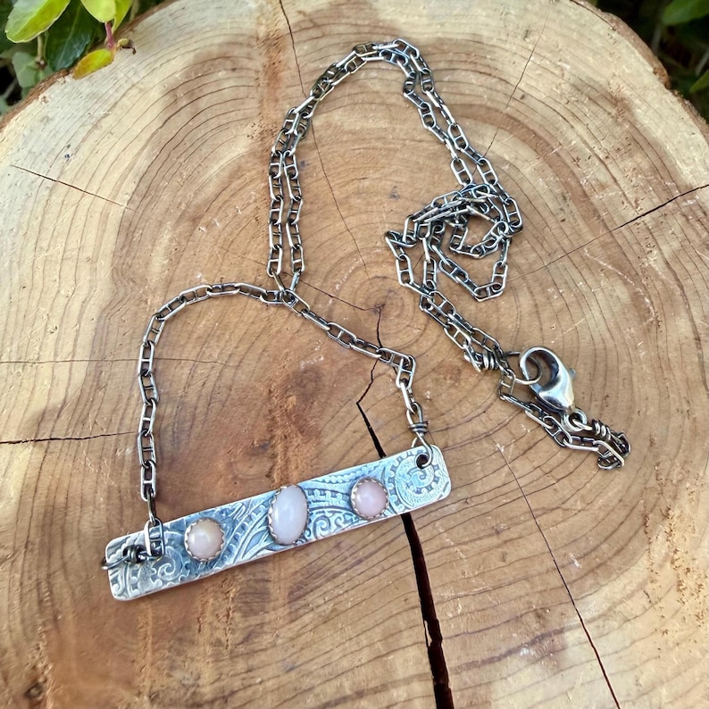 May include: A silver-toned metal bar necklace featuring three oval-shaped pink stones set in a row. The rectangular bar has an embossed swirling pattern and is attached to a silver-toned chain with a lobster claw clasp closure.
