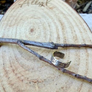 May include: A silver-colored ring with an engraved leaf pattern. The ring is resting on a small tree branch, set against a wooden background. The word "Allyk" is written on the wood.