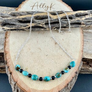 May include: A silver chain necklace with turquoise and black beads. The turquoise beads have brown matrix patterns. The necklace is displayed on a wooden surface with the handwritten text "Allyk".