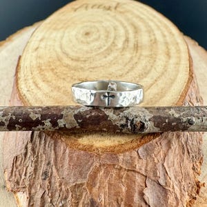 May include: A silver ring with a hammered texture and a black cross design. The ring is displayed on a wooden branch, showcasing its unique craftsmanship and religious theme. The ring's design is simple yet elegant.