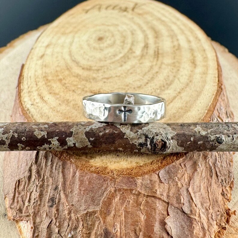 May include: A silver ring with a hammered texture and a black cross design. The ring is displayed on a wooden branch, showcasing its unique craftsmanship and religious theme. The ring's design is simple yet elegant.