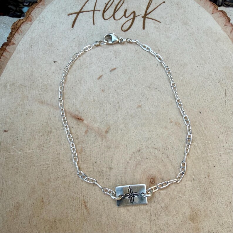 May include: A silver chain bracelet with a rectangular pendant. The pendant features a cross design. The bracelet is displayed on a wooden surface. The word "Allyk" is written in cursive at the top of the image.
