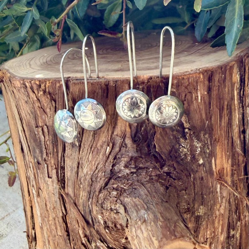 K&ouml;nnte beinhalten: Zwei Paar Ohrh&auml;nger aus silberfarbenem Metall mit kreisf&ouml;rmigen Scheiben, die gravierte Sonnenstrahlen- und Blumenmuster aufweisen. Jeder Ohrring hat einen langen, gebogenen Haken zur Aufh&auml;ngung und zeigt feine, ge&auml;tzte Details auf den Oberfl&auml;chen.