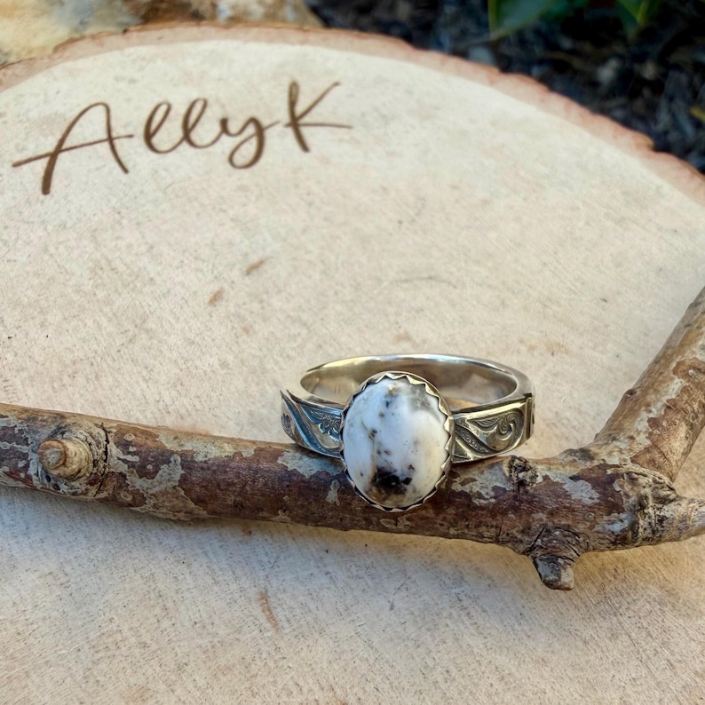 May include: A silver ring with an oval, white stone with black and brown speckles. The ring has a decorative band with etched details. The ring is displayed on a small, brown branch.