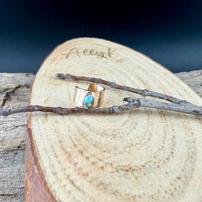 May include: A gold-colored ring with a turquoise stone. The ring is set on a wooden surface with a branch. The ring has a textured band and an oval-shaped turquoise stone. The background is dark.