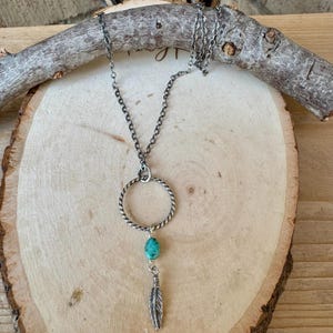 May include: A silver-toned necklace with a turquoise stone and feather pendant. The necklace features a circular pendant with a twisted design. The pendant hangs from a chain, resting on a wooden surface.