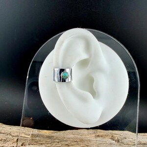 May include: A silver ear cuff with a turquoise stone. The cuff is shown on a white ear model against a black background. The cuff has a textured, horizontal striped design.