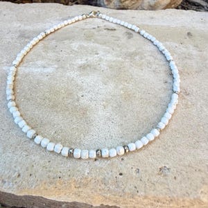 May include: White beaded necklace with silver accents. The necklace features small, square-shaped white beads strung together with silver-colored metal spacers. The clasp is silver. The necklace is displayed on a light-colored stone surface.