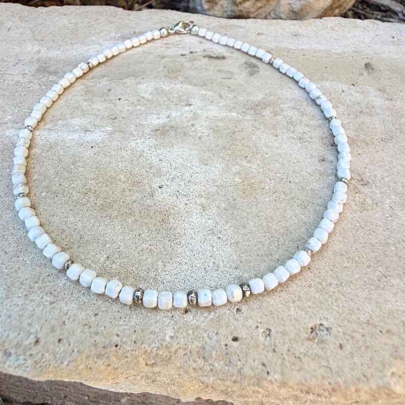 May include: White beaded necklace with silver accents. The necklace features small, square-shaped white beads strung together with silver-colored metal spacers. The clasp is silver. The necklace is displayed on a light-colored stone surface.