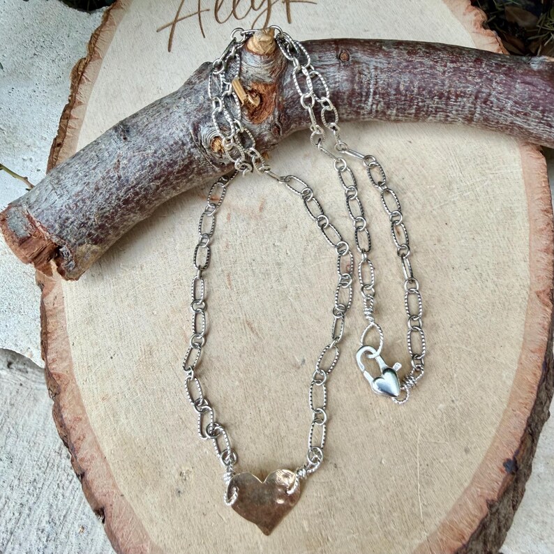 May include: A silver-toned chain necklace featuring a textured oval link design and a single heart-shaped pendant at the center. The necklace includes a heart-shaped lobster claw clasp closure, providing a secure and functional fastening mechanism.