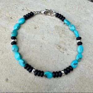 May include: A beaded bracelet featuring turquoise oval beads and black round beads, interspersed with silver-toned metal accents. The bracelet is secured with a silver clasp.