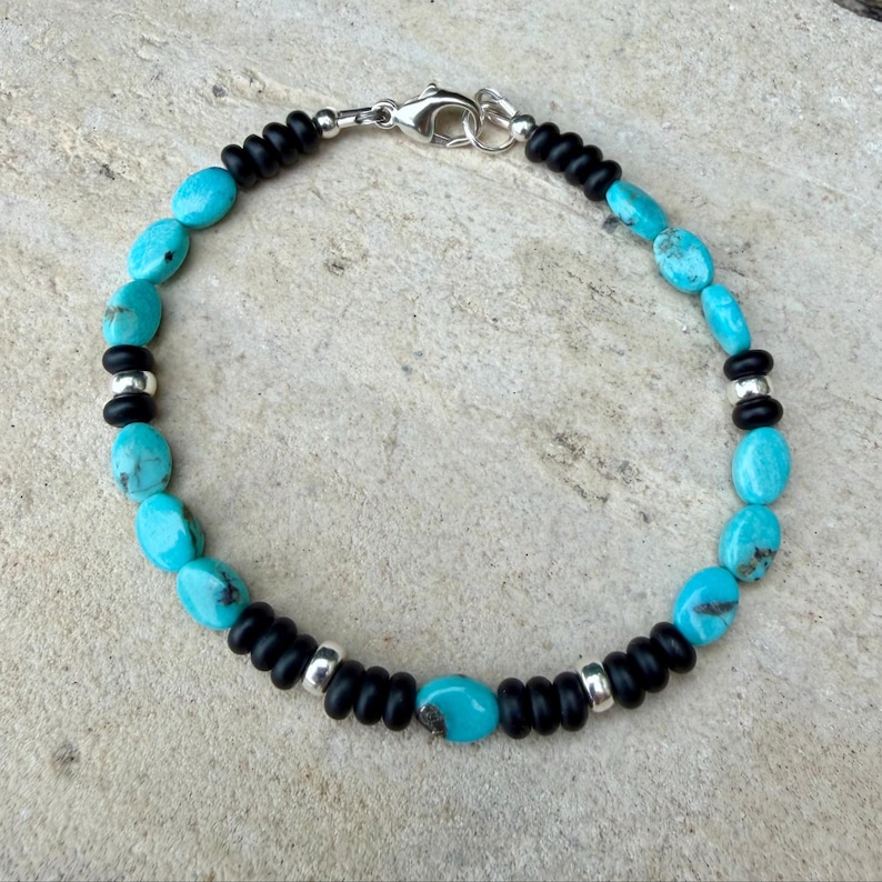 May include: A beaded bracelet featuring turquoise oval beads and black round beads, interspersed with silver-toned metal accents. The bracelet is secured with a silver clasp.
