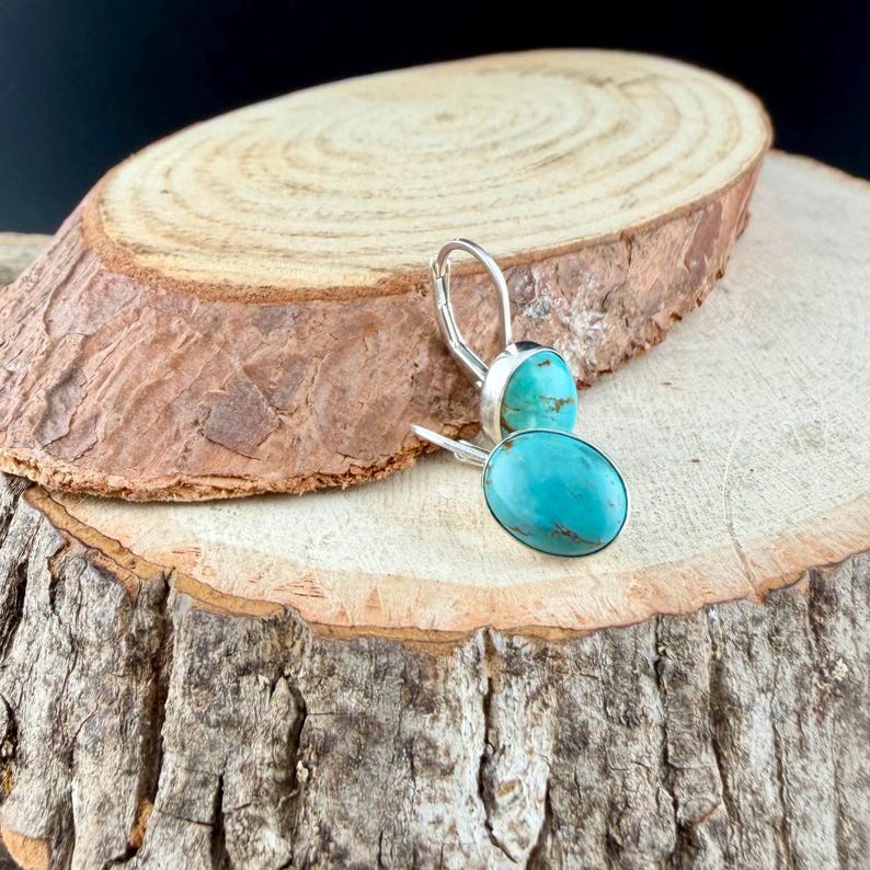 May include: A pair of silver leverback earrings featuring oval turquoise stones. The earrings are displayed on a rustic wooden surface, showcasing the vibrant blue-green color of the gemstones. The earrings have a simple, elegant design.