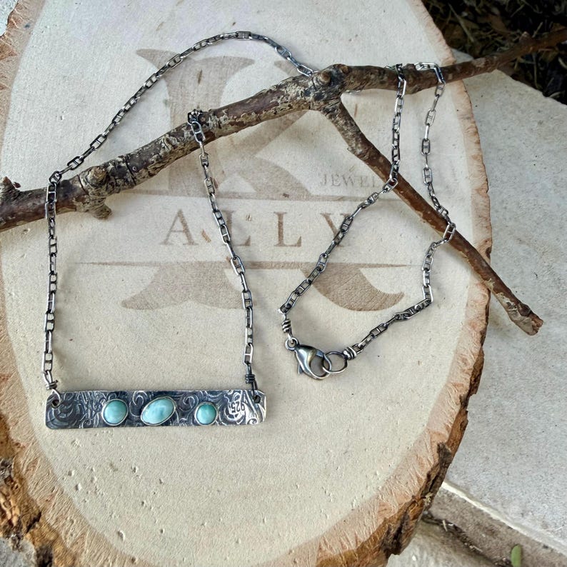 May include: A silver necklace featuring a rectangular bar pendant with an oxidized, textured surface and three oval, light blue stones. The pendant is attached to a silver chain with a lobster clasp. The metal is stamped with 925.