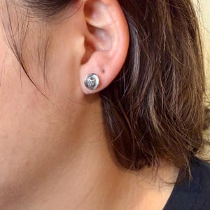 May include: A silver stud earring with a circular design is shown on an ear. The earring has a textured surface and a small, detailed pattern in the center. The background is blurred, focusing attention on the jewelry.