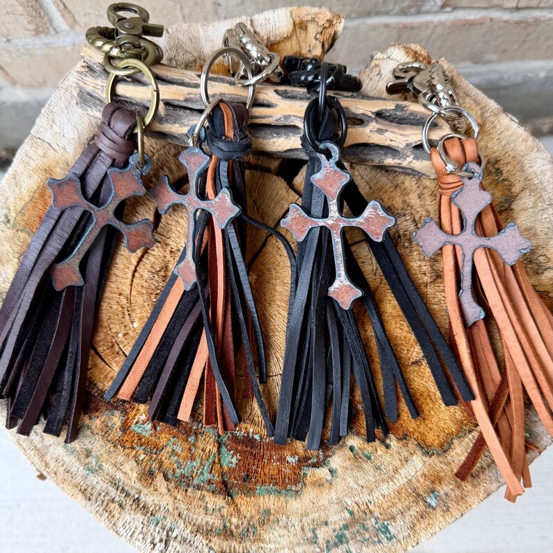Op de afbeelding: Vier sleutelhangers met leren kwastjes en metalen kruisbedels. Elke sleutelhanger is voorzien van een draaibare karabijnhaak en een sleutelring. De kwastjes zijn gemaakt van donkerbruin, zwart en lichtbruin leer met verweerde kruisjes.