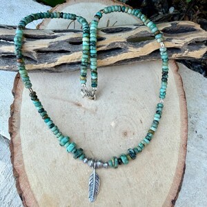 May include: Turquoise beaded necklace with a silver feather pendant. The necklace features small, round turquoise beads in shades of green and blue, strung together with silver accents. The pendant is a detailed silver feather.