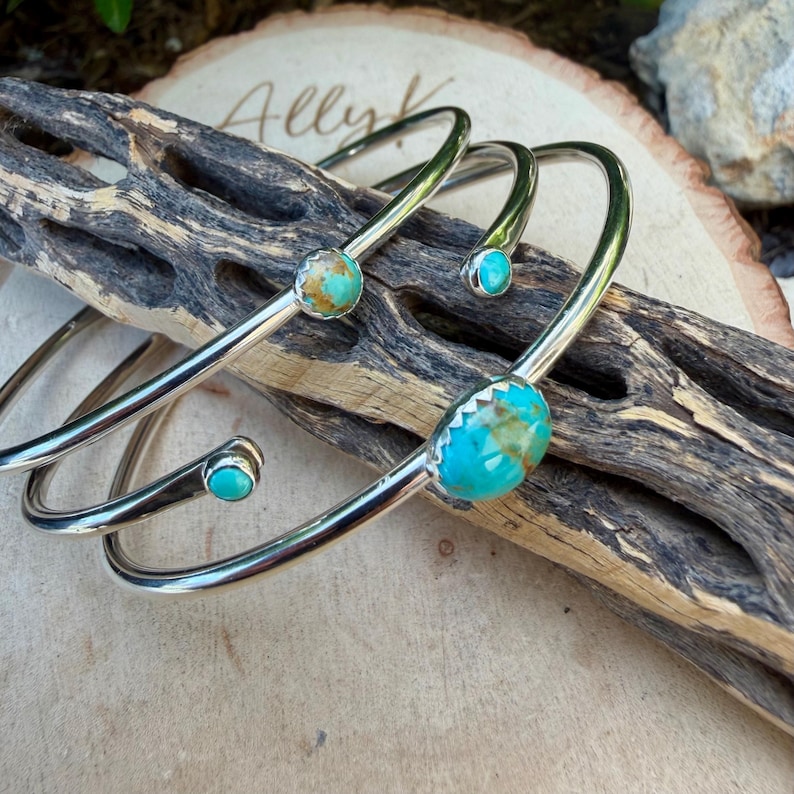 May include: Silver-toned metal cuff bracelets with turquoise stones. The bracelets have a simple, elegant design, with the turquoise stones set in the center and at the ends of the curved bands. The bracelets are displayed on a piece of wood.