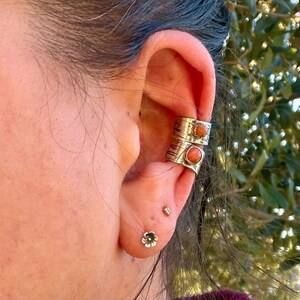 May include: Two silver-toned ear cuffs featuring textured bands and round, orange-colored stones, worn on the upper cartilage of the ear, accompanied by a small silver-toned flower-shaped stud earring in the lower earlobe.