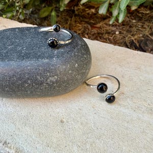 May include: Two open-style rings crafted from silver-toned metal, each featuring two round, polished black stones at the ends. The bands have a textured, hammered finish, and the design allows for an adjustable fit on the finger.