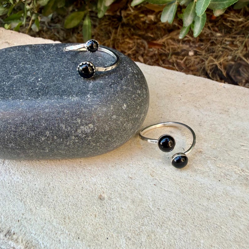 May include: Two open-style rings crafted from silver-toned metal, each featuring two round, polished black stones at the ends. The bands have a textured, hammered finish, and the design allows for an adjustable fit on the finger.