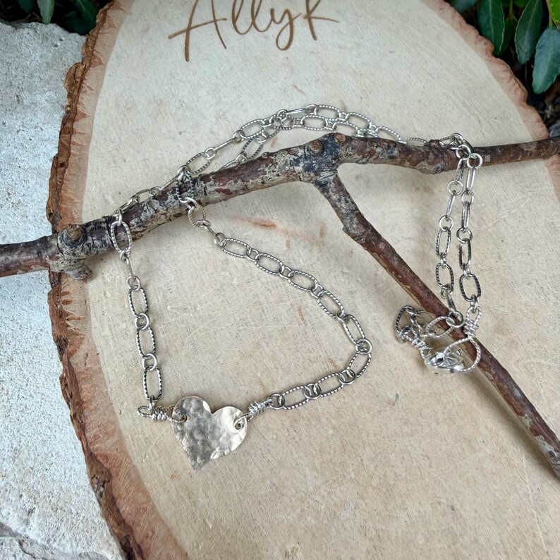 May include: A silver-toned necklace featuring a hammered metal heart pendant attached to a textured oval link chain. The chain is draped over a brown wooden branch, showcasing the metallic finish and the irregular, dimpled surface of the heart.