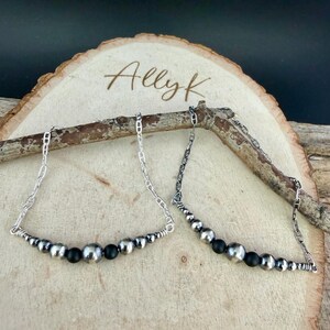 May include: Two silver necklaces with a chain design and a row of silver and black beads. The necklaces are displayed on a wooden slice with the text "Ally K" in cursive.