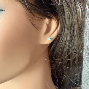 May include: A close-up of a person's ear with a small, round, light blue stud earring. The earring is set in a silver or white metal. The person has brown hair.