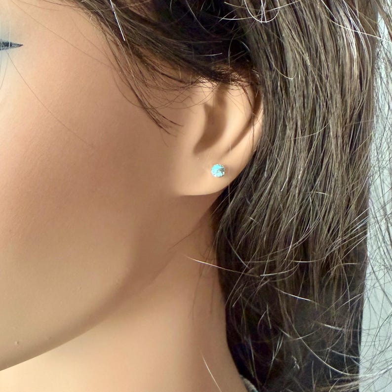 May include: A close-up of a person's ear with a small, round, light blue stud earring. The earring is set in a silver or white metal. The person has brown hair.
