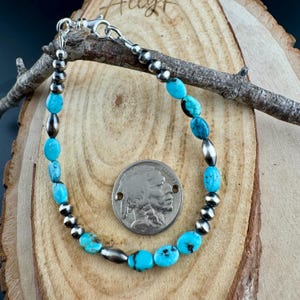 May include: A turquoise and silver beaded bracelet with a lobster clasp. The bracelet features oval turquoise beads and silver-colored round and oblong beads. A silver coin is next to the bracelet.