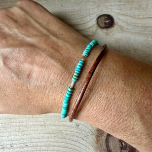 May include: A two-piece bracelet set consisting of one strand of small, round, turquoise-colored beads with silver-toned accents and one plain, smooth, dark brown leather cord bracelet worn together on the wrist.