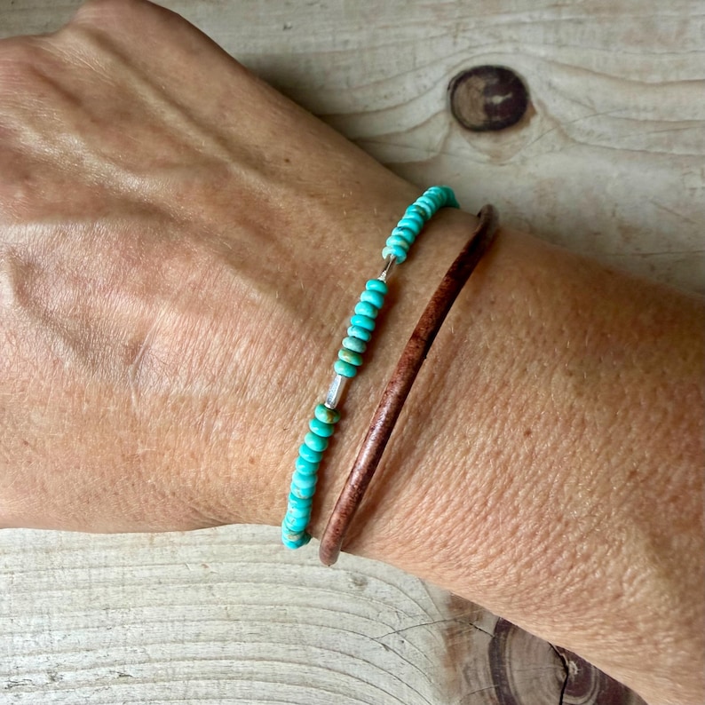 May include: A two-piece bracelet set consisting of one strand of small, round, turquoise-colored beads with silver-toned accents and one plain, smooth, dark brown leather cord bracelet worn together on the wrist.