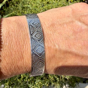 May include: A silver-toned cuff bracelet featuring a repeating geometric pattern of etched triangles and diamond shapes. The metal surface has a polished finish with dark recessed lines that highlight the intricate, symmetrical engraved design.